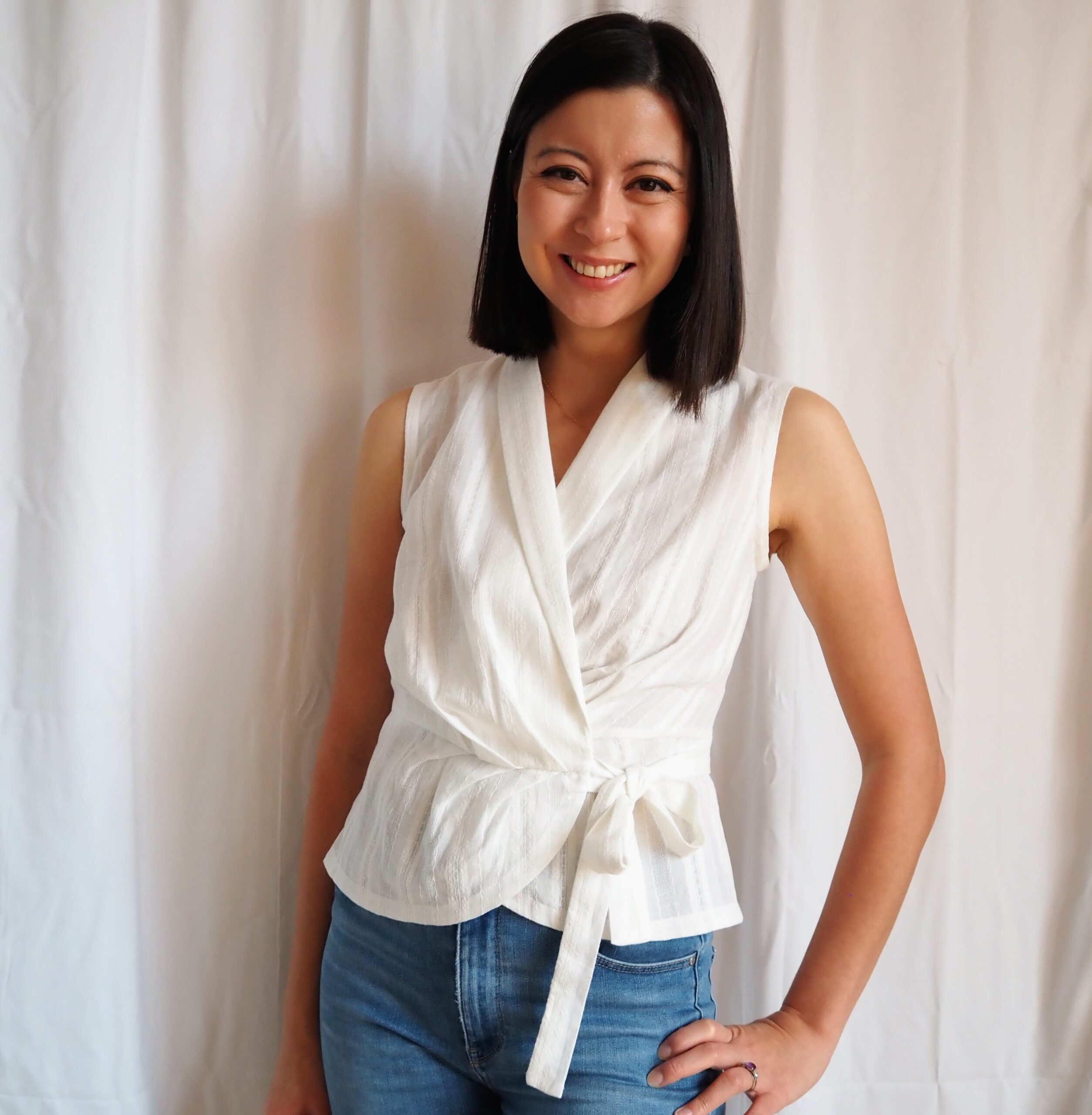 A half white, half Asian woman stands smiling in front of a white curtain. She is wearing a white sleeveless wrap blouse and blue jeans. 